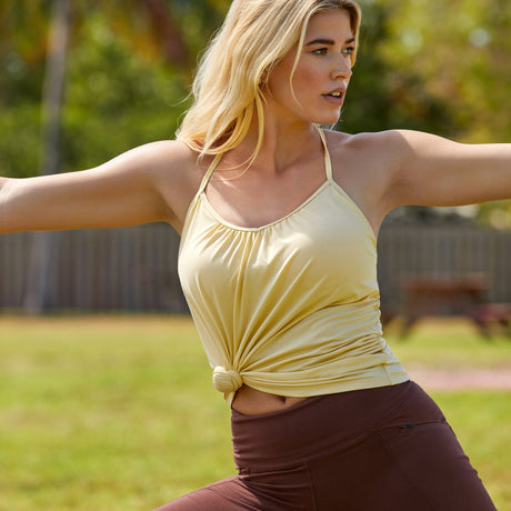 A person performs a yoga pose outdoors, wearing a yellow sleeveless top knotted at the waist and brown leggings. They stand on one leg with arms extended, against a backdrop of grass, a wooden fence, and softly focused trees.