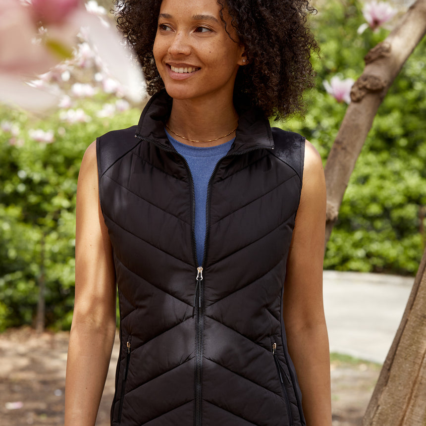 Woman wearing the Free Country Women's Quilted Hybrid Vest in black, featuring a unique quilted design with stretch knit panels and REPREVE® synthetic fill, standing outdoors near blooming trees.