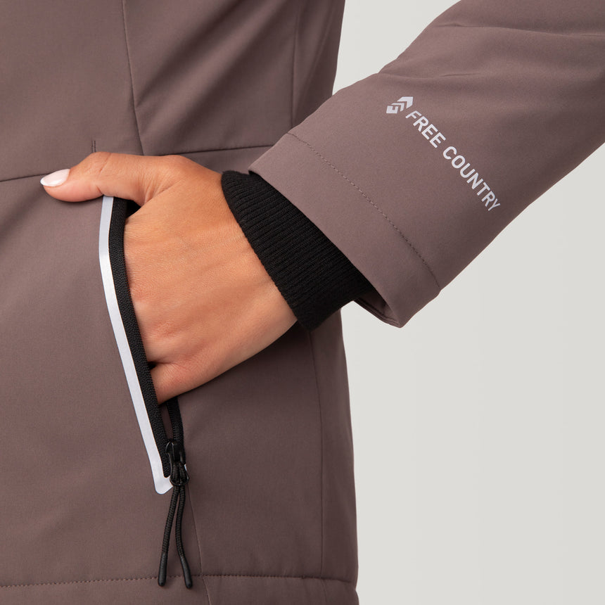 Woman wearing Free Country Women's FreeCycle® Thermo Super Softshell® II Jacket in light brown with a full front zipper opening, lower pockets, soft lining, a detachable hood with an adjustable drawcord, and water resistant and wind resistant stretch performance fabric, light grey background. Best women’s jacket for winter weather.