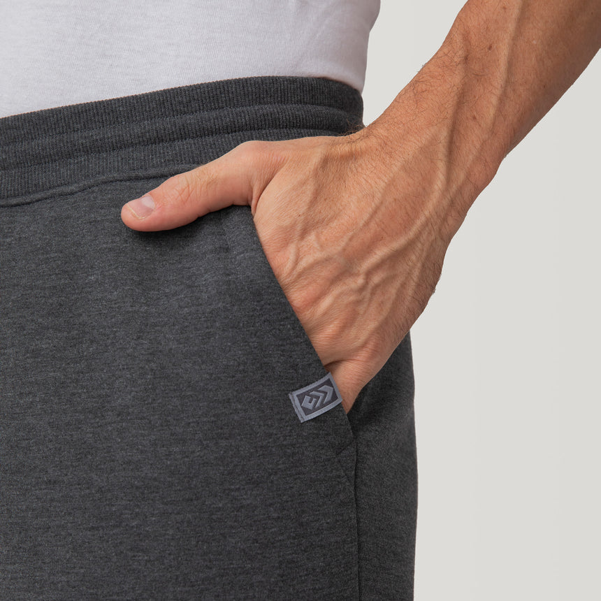 A close-up shows a person wearing Free Country Men’s Sherpa Lined Joggers in dark gray with a white shirt. Their right hand is in the pocket, and a small logo patch near the pocket highlights trendy athleisure style.
