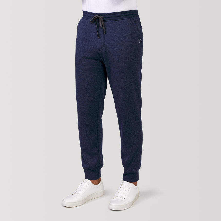 A man in Free Country Men’s Sherpa Lined Jogger, Navy Heather, with a drawstring waist and white sneakers stands against a light grey background—ideal for cozy athleisure style.