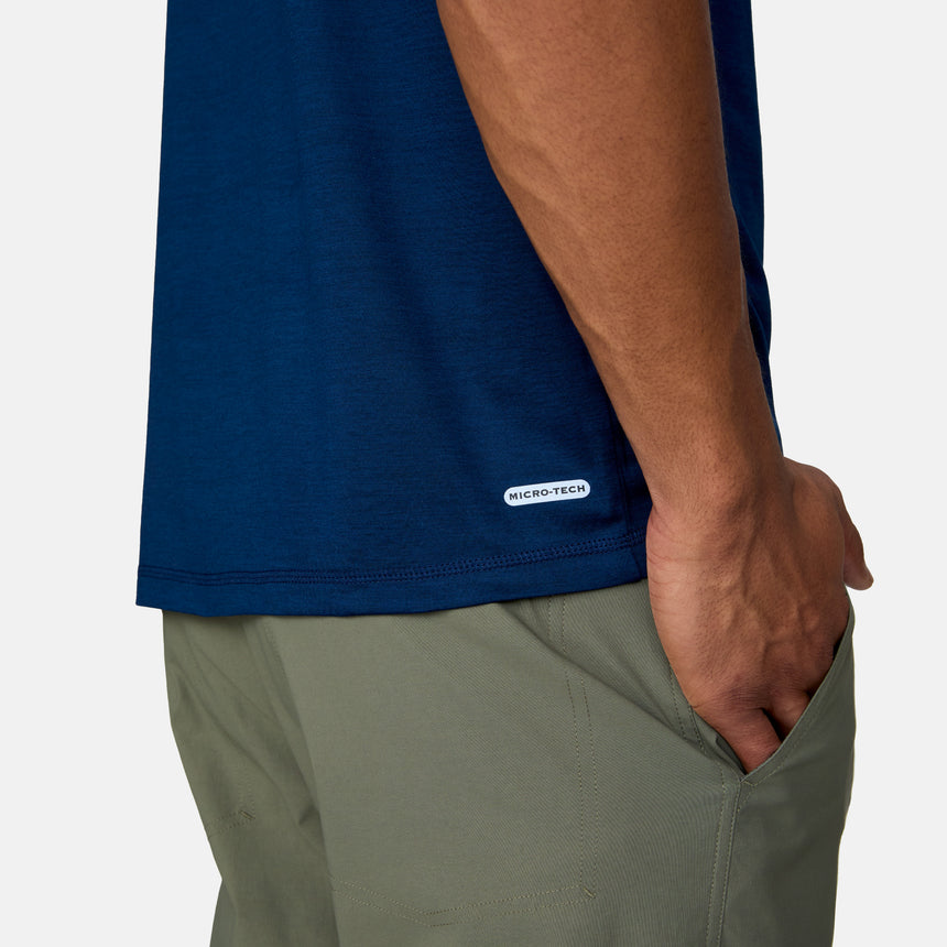 A person in an untucked dark blue Men's Super Soft Short Sleeve Crew Tee by Clearance and olive green pants stands with one hand in their pocket. The ultra-soft tee features a small MICRO-TECH label by the hem for all-day comfort.