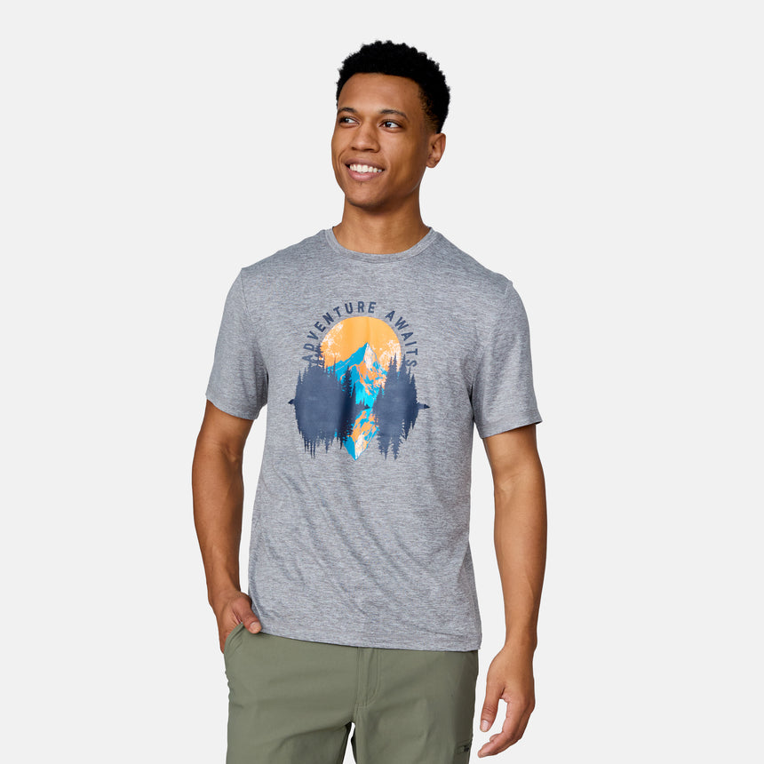 A smiling man with short, curly hair wears the Clearance Men's Super Soft Short Sleeve Crew Tee - 2 Pack in gray, featuring mountains, trees, a sunset, and "Adventure Awaits," paired with olive-green pants and one hand in his pocket.