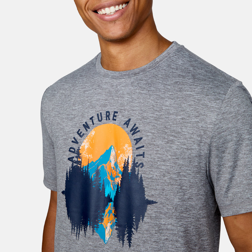 A person in a Clearance Men's Super Soft Short Sleeve Crew Tee - 2 Pack, gray with a vibrant mountains, trees, and sun graphic and "ADVENTURE AWAITS" text. This moisture-wicking tee keeps them smiling and comfy all day.