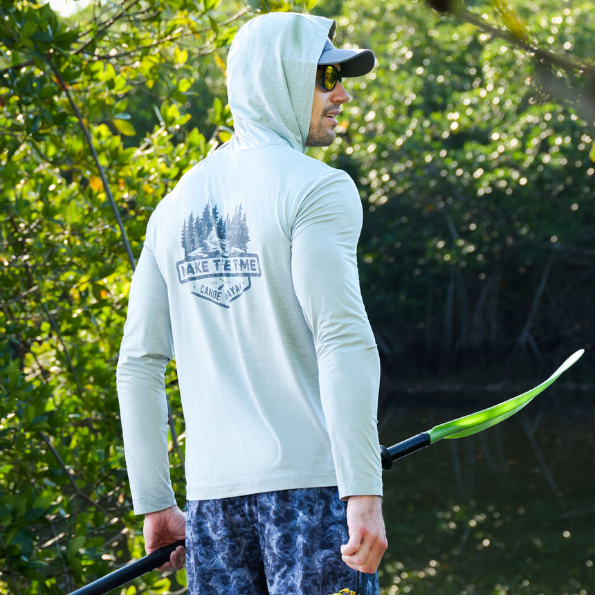 A man wearing the Free Country Men's Microtech® Hooded Graphic Sunshirt with UPF 50+ holds a green-bladed paddle near water. The shirt says "Lake Time Canoe & Kayak" with pine trees, and there are trees in the background.