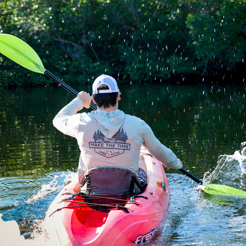 Wearing a Free Country Men's Microtech® Hooded Graphic Sunshirt (UPF 50+), a person in a cap paddles a red kayak through calm water, surrounded by green trees. Water splashes from the bright green paddle. The hoodie reads MAKE THE TIME CAMP & CANAL.