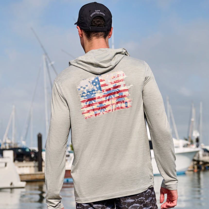 Man wearing Free Country Men's Microtech® Hooded Graphic Sunshirt in Iron, size M, made from lightweight four way stretch performance fabric featuring upf, moisture wicking, and anti-odor technology, styled with fish printed swim shorts at the marina #color_iron
