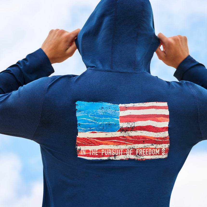 Man wearing Free Country Men's Microtech® Hooded Graphic Sunshirt in Trails, size M, made from lightweight four way stretch performance fabric featuring an american flag design and upf, moisture wicking, and anti-odor technology, styled with navy shorts #color_trails