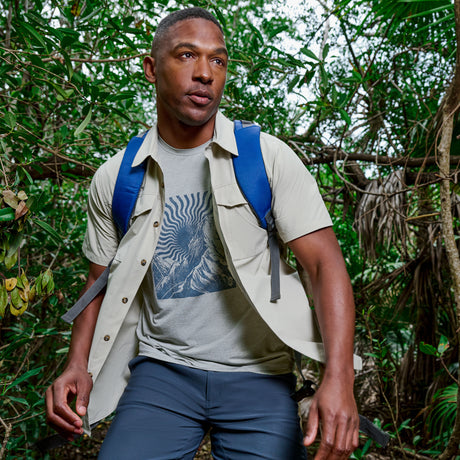 A man wearing the Free Country Men's Super Soft Microtech® Short Sleeve Graphic Crew Tee, blue backpack, and dark pants walks through a lush green forest, looking focused and ready for adventure in comfort and style.