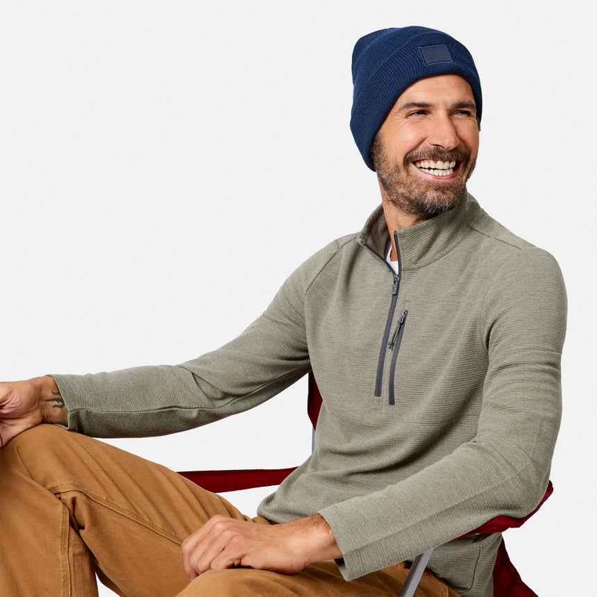 A man with a beard smiles while sitting in a chair, wearing the Free Country Men’s Altitude Quilt 1/2 Zip Mock Neck Shirt in olive green, along with a blue beanie and tan pants. The background is plain and light-colored.