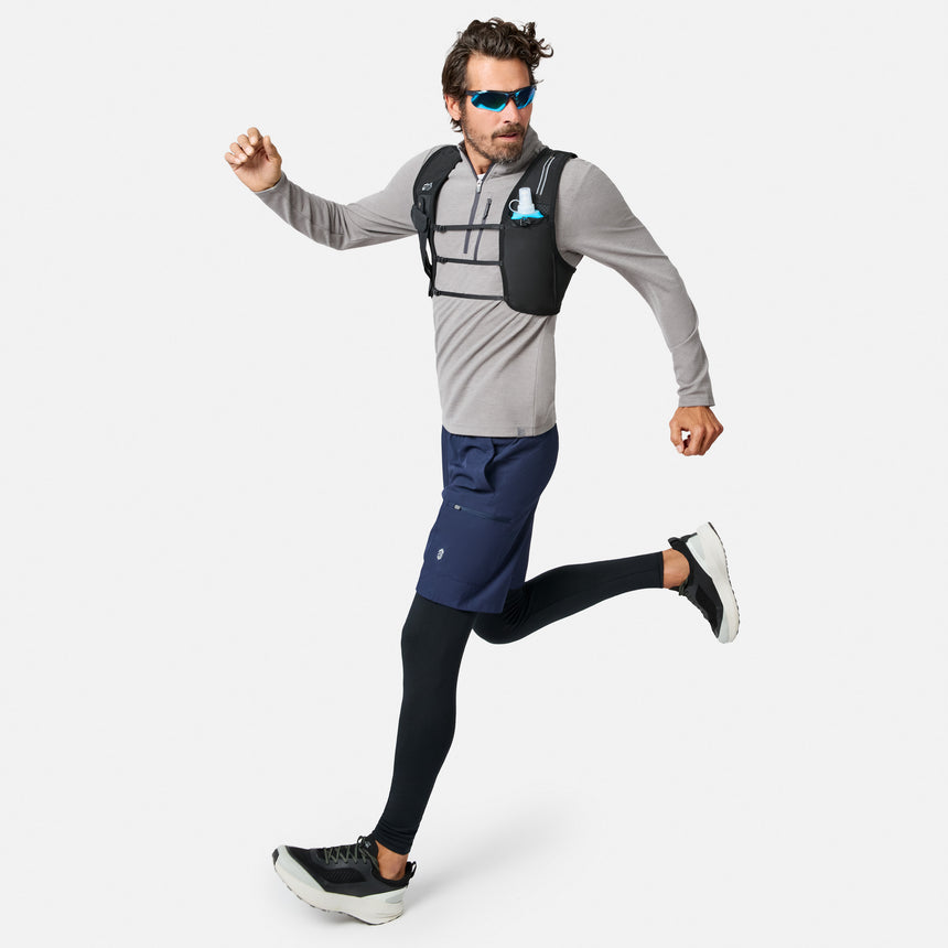 A man wearing sunglasses, the Free Country Men’s Altitude Quilt 1/2 Zip Mock Neck Shirt in gray, navy shorts, black leggings, and running shoes is running. He has on a black hydration vest and carries a collapsible blue water bottle in a chest pocket.