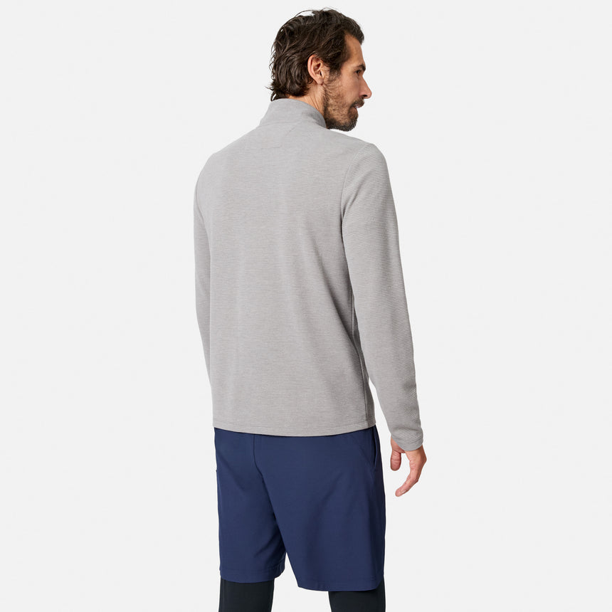 A man with dark hair is standing and facing away from the camera against a plain white background, wearing the Free Country Men’s Altitude Quilt 1/2 Zip Mock Neck Shirt in light gray and navy shorts.