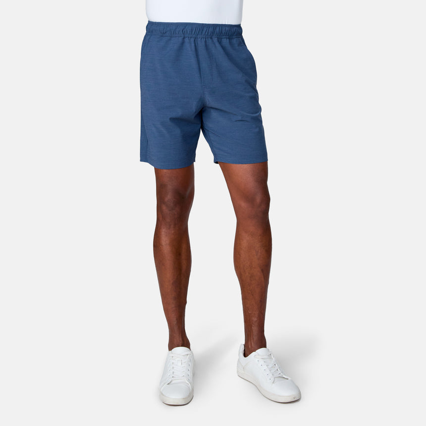 A person wearing Clearance Men's Trek Short in blue stands against a plain light gray background. The image is cropped from the waist down, highlighting the four-way stretch shorts and legs, paired with white sneakers.