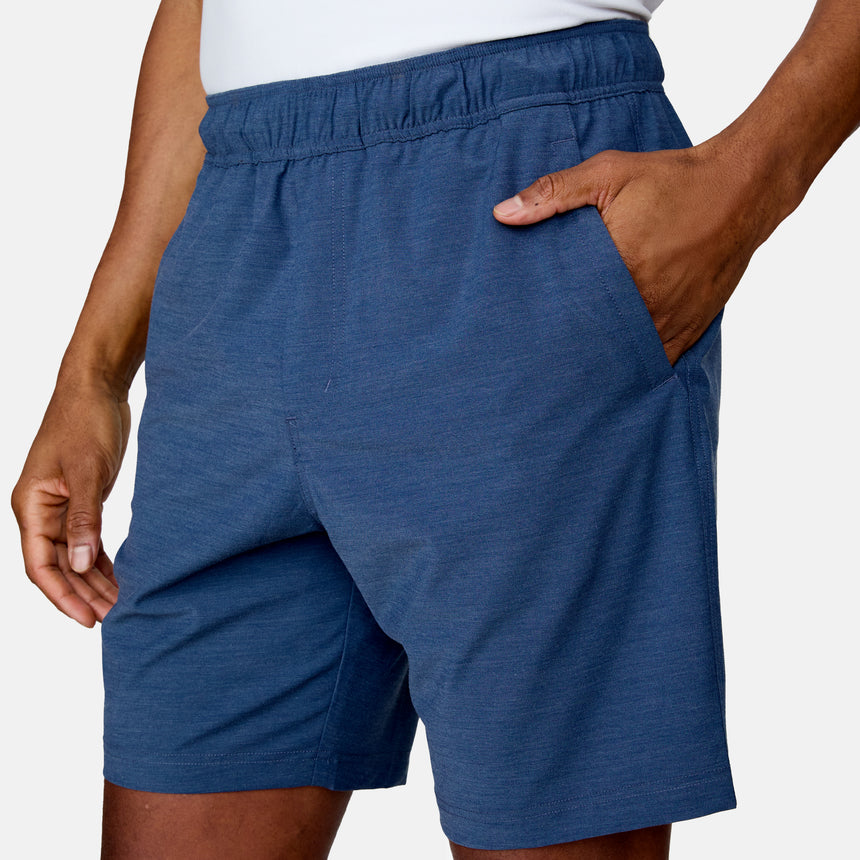 A person wearing Clearance Men's Trek Shorts in blue, featuring lightweight four-way stretch fabric and an elastic waist, with a white shirt and one hand in a pocket, stands against a plain background.