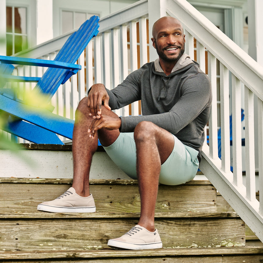 Man wearing the Free Country Men's Boardwalk Woven Pull-On Trek Short in Basil, size M, made from lightweight breathable polyester fabric and featuring an elastic waistband and slanted pockets, styled with a grey hoodie on a front porch #color_ basil