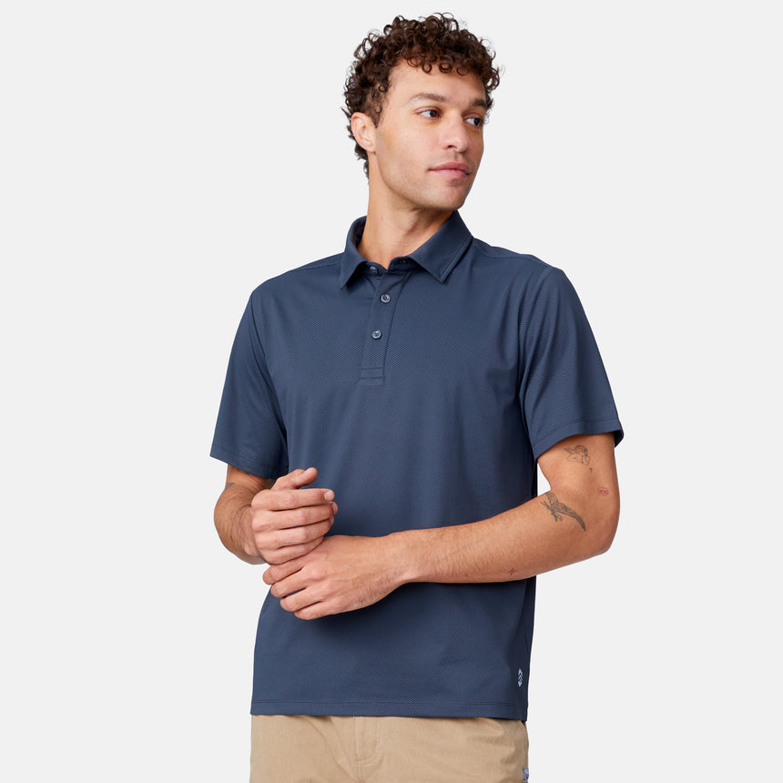 A man with curly hair models the Clearance Men's Tech Pique Polo Shirt in navy blue and beige pants, standing against a plain white background, looking slightly left with his hands together.