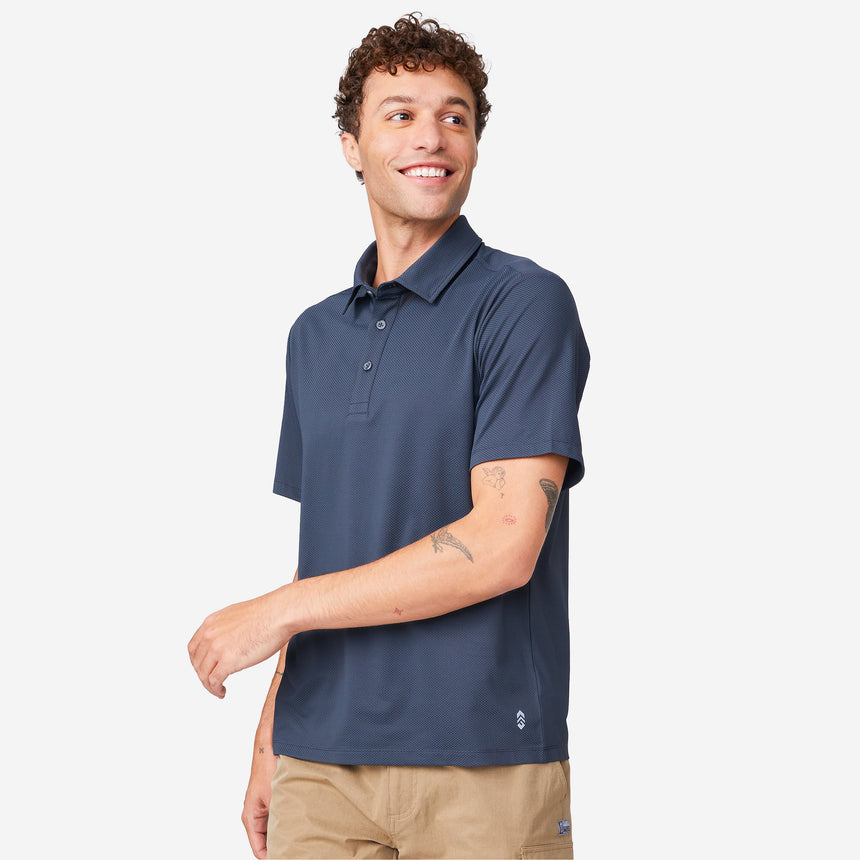 A person in a dark blue Men's Tech Pique Polo Shirt by Clearance and tan shorts smiles and looks to the side against a plain white background.