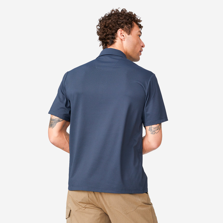 A man with curly hair is seen from behind wearing a Clearance Men's Tech Pique Polo Shirt in navy and tan shorts, standing against a plain white background.