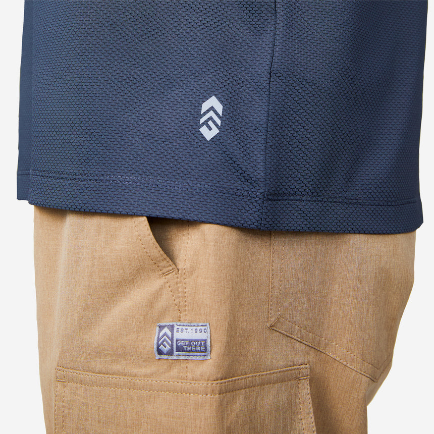 A close-up of a person wearing the Men’s Tech Pique Polo Shirt in navy blue by Clearance and tan pants, both featuring small white and blue stitched logo patches, set against a plain white background.