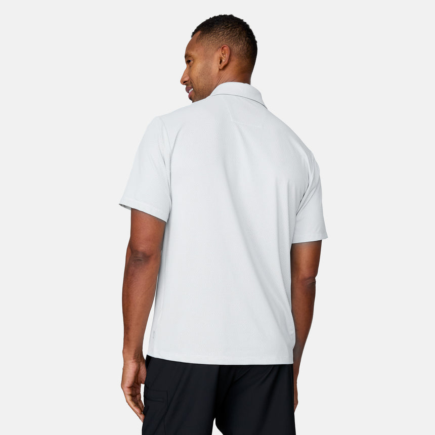 A man faces away, glancing over his shoulder in a plain light setting. He wears the Clearance Men's Tech Pique Polo Shirt in light gray and black shorts.