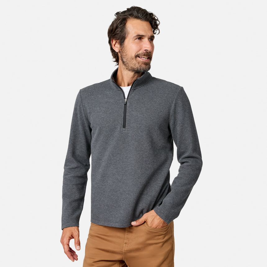 A man with wavy brown hair and a beard is wearing the Free Country Men’s Twill Interlock 1/4 Zip Long Sleeve Shirt in a dark gray shade, paired with tan pants. Crafted from breathable knit fabric, the pullover features a sleek quarter zip and mock neck design. He stands against a plain white background, looking to his left with his right hand in his pocket.