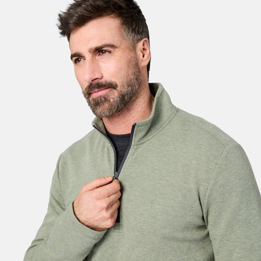 A man with short brown hair and a beard wears the Free Country Men’s Twill Interlock 1/4 Zip Long Sleeve Shirt in a light green color over a dark shirt. He is looking slightly to the side while pulling up the zipper, with a plain, light-colored background behind him.