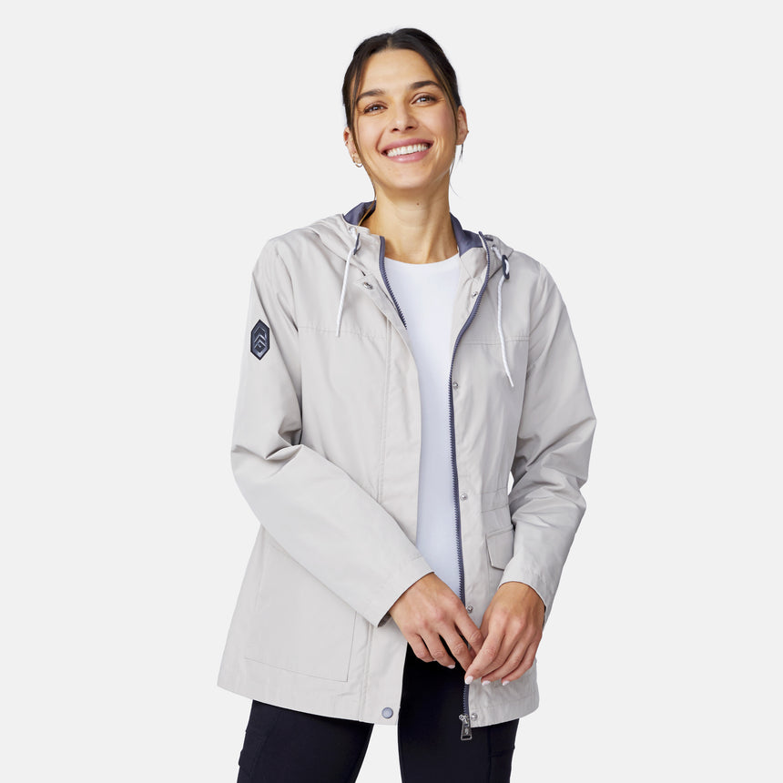 A smiling woman with dark hair tied back wears the Free Country Women's Rain Away Anorak Rain Jacket in light gray over a white shirt, standing against a plain white background and holding the front of her jacket with both hands.