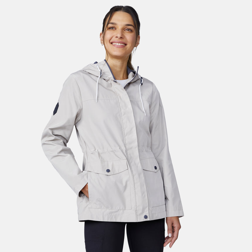 A woman in the Free Country Women's Rain Away Anorak Rain Jacket, light gray with an adjustable hood and pockets, smiles with her hands in the pockets while looking slightly up against a plain white background.