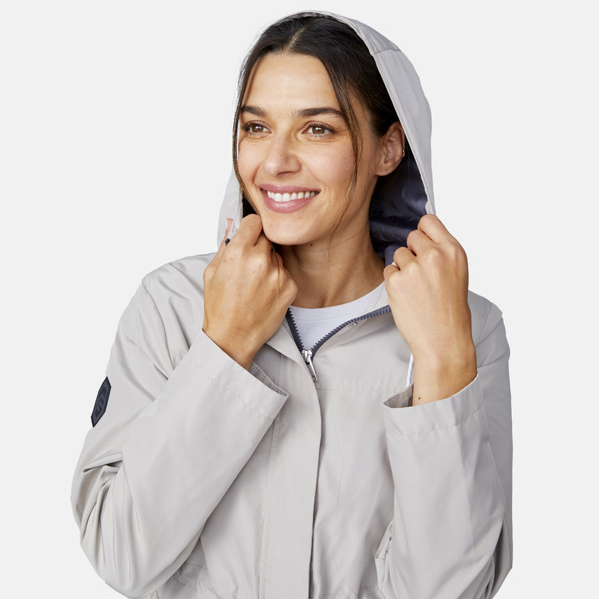 A woman smiles and looks to the side, holding the adjustable hood of her light gray Free Country Women's Rain Away Anorak Rain Jacket against a plain light background.