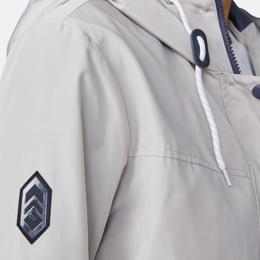 Close-up of the Free Country Women's Rain Away Anorak Rain Jacket in light beige, featuring a white drawstring hood, black geometric sleeve patch, and partially visible zipper and snap button details.