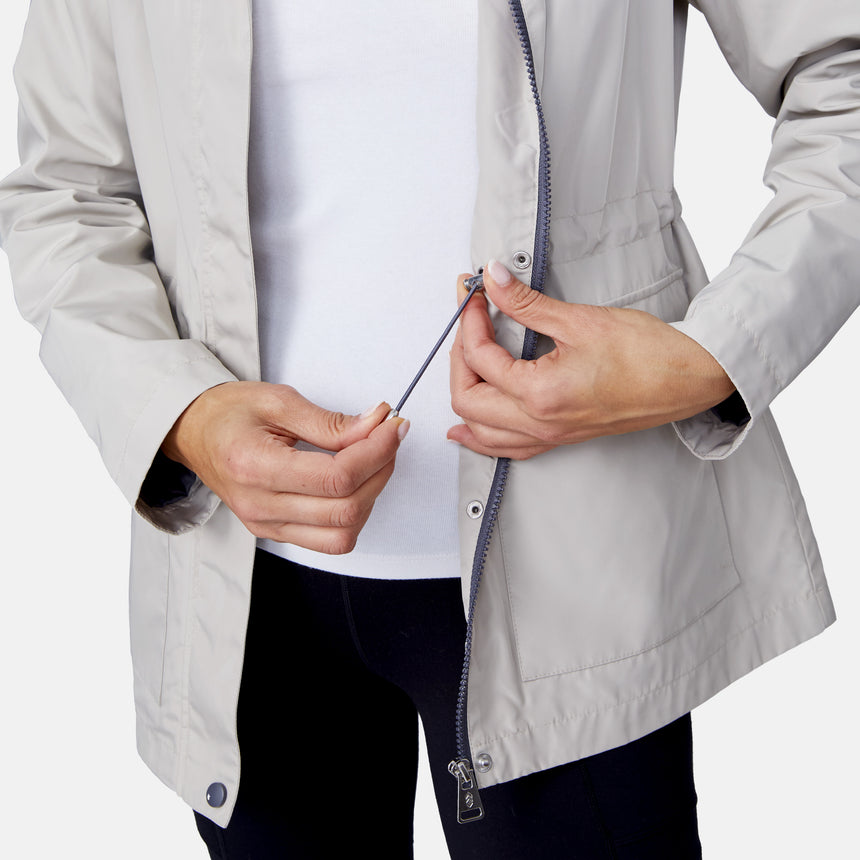 A person adjusts the elastic drawstring inside a partially unzipped Women's Rain Away Anorak Rain Jacket by Free Country, revealing a white shirt underneath. Only their torso and hands are visible.