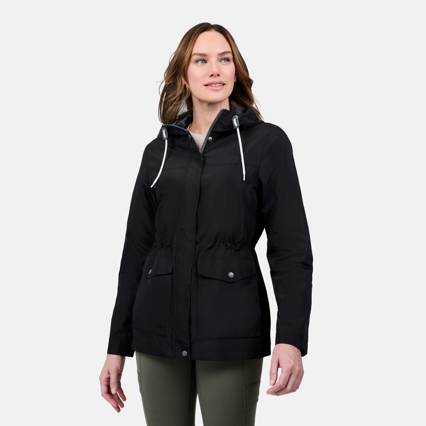 A woman with light brown hair smiles while wearing the Free Country Women's Rain Away Anorak Rain Jacket in black and olive green pants, standing against a plain light gray background. The jacket features water resistance and an adjustable hood.