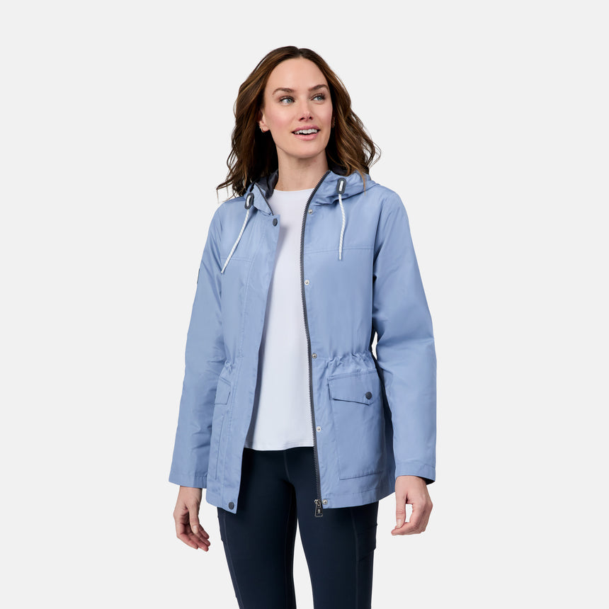 A woman smiles and looks to the side while wearing the Free Country Women's Rain Away Anorak Rain Jacket in light blue over a white shirt and dark pants, standing against a plain white background.