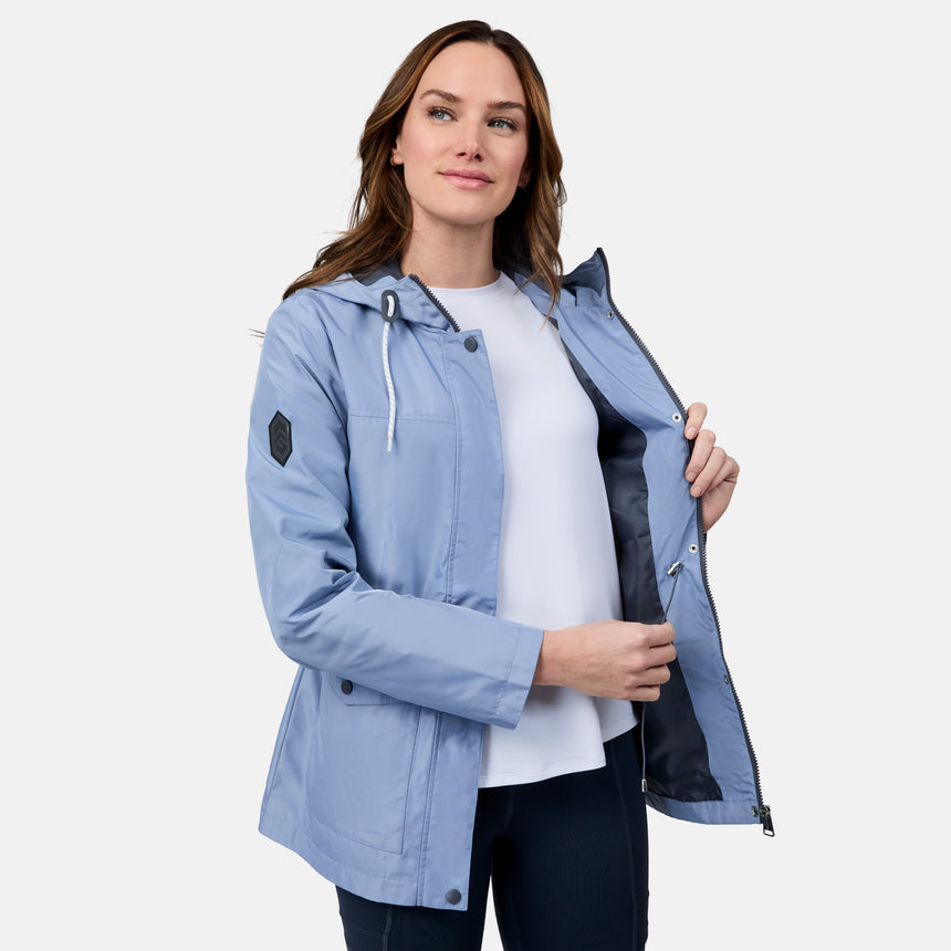A woman wears the Free Country Women's Rain Away Anorak Rain Jacket, a light blue water-resistant style with an adjustable hood, over a white shirt against a plain background, holding the jacket open and smiling slightly.