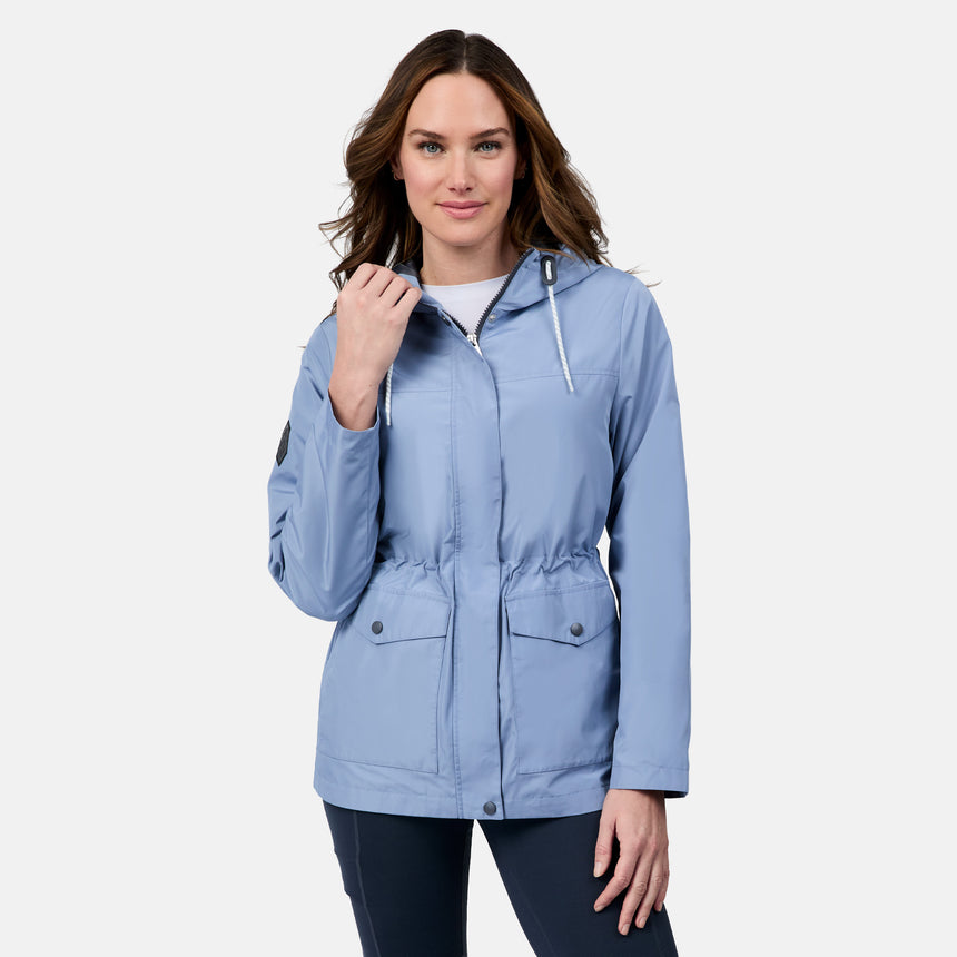 A woman with long brown hair smiles in the Free Country Women's Rain Away Anorak Rain Jacket—a light blue, water-resistant coat with adjustable hood, front zipper, and snap buttons—standing against a plain white background.