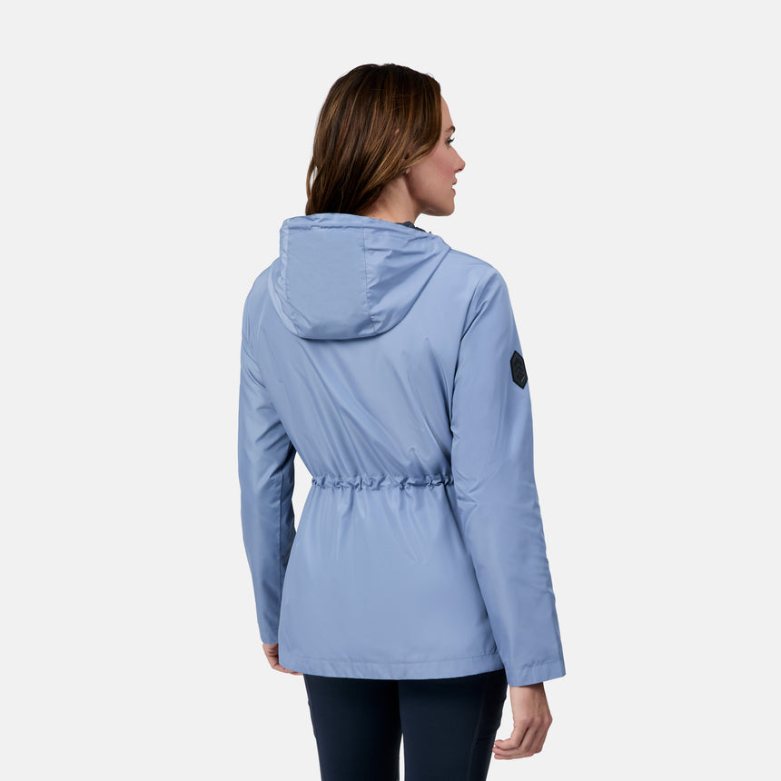 A woman with light brown hair is seen from the back wearing the Free Country Women's Rain Away Anorak Rain Jacket in light blue, featuring a water-resistant finish, adjustable hood, fitted waist, left sleeve patch, and paired with dark leggings against a white background.