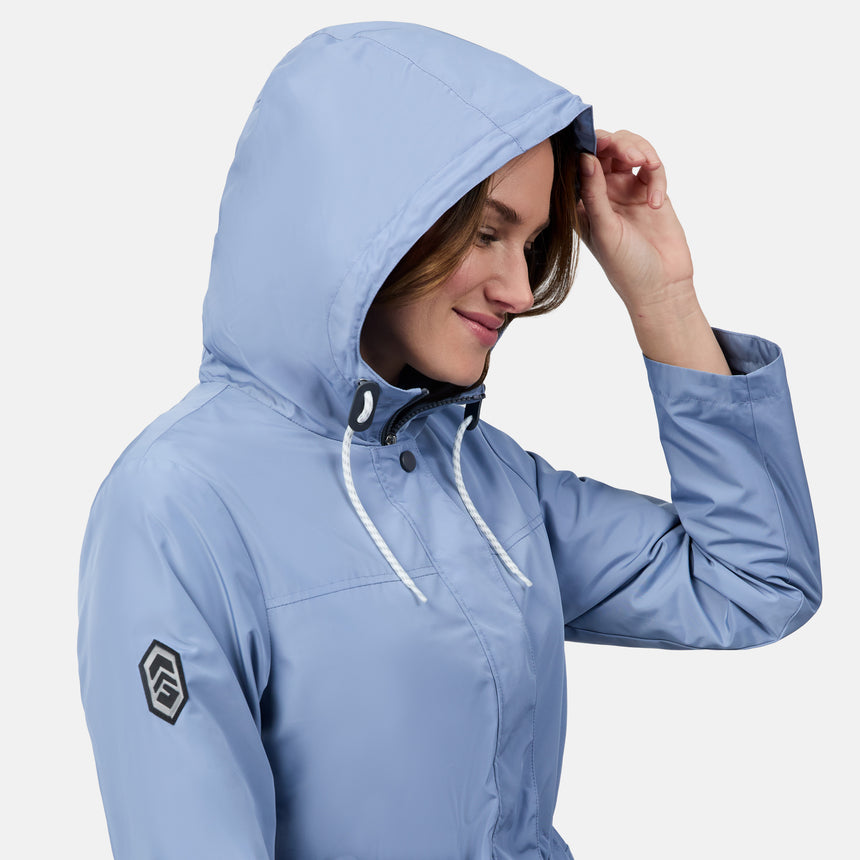 A woman smiles, adjusting the hood of her light blue Free Country Women's Rain Away Anorak Rain Jacket. The water-resistant jacket features white drawstrings and a black logo patch on the sleeve against a plain white background.