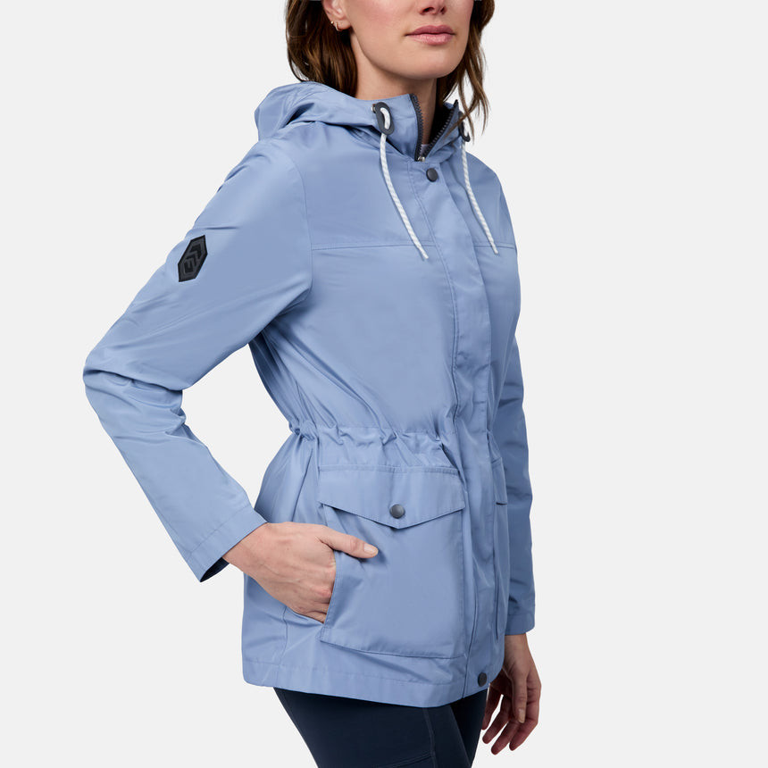 A woman wears the Free Country Women's Rain Away Anorak Rain Jacket, a light blue, water-resistant style featuring an adjustable hood, drawstrings, and flap pockets, standing with one hand in her pocket against a plain background.