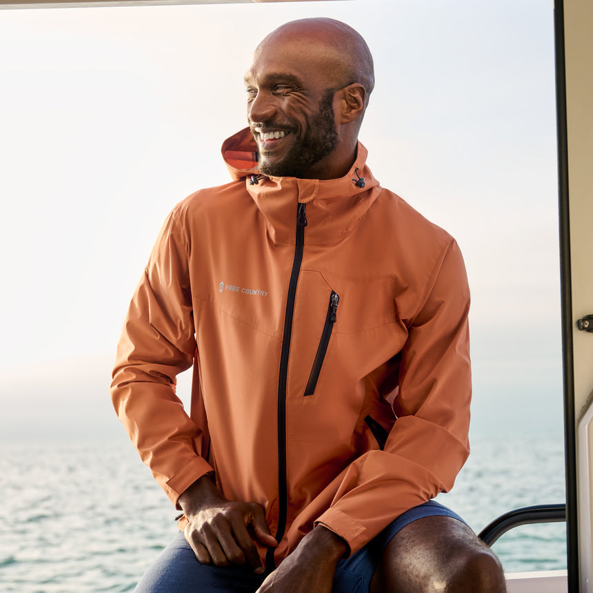 Man wearing Free Country Men's Hydro Lite X2O Status Jacket in Autumn, size M, made from lightweight four way stretch water-resistant performance fabric featuring tab cuffs, an attached hood with a drawcord, and a chest and lower pockets, styled with blue shorts #color_autumn