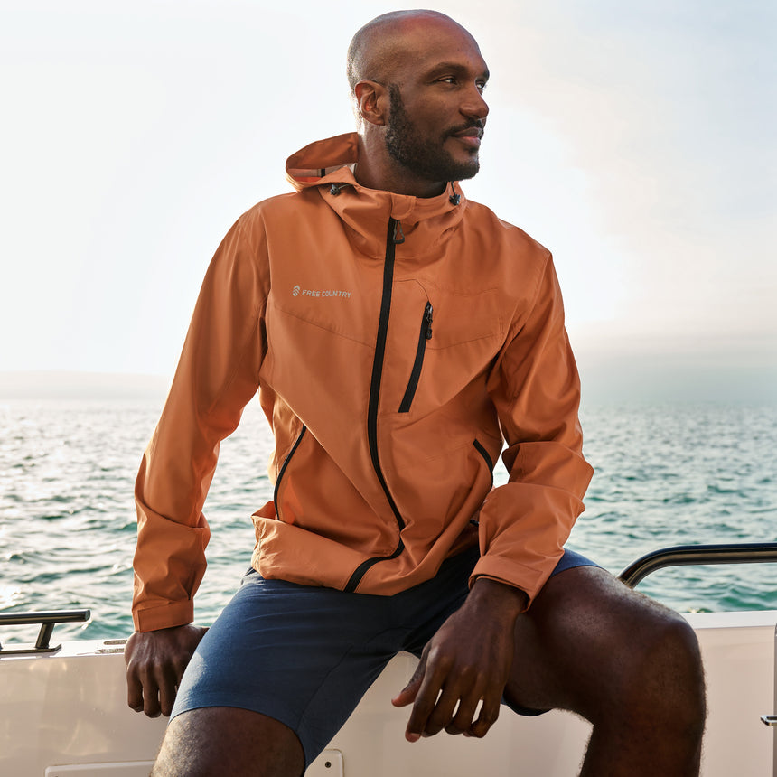 Man wearing Free Country Men's Hydro Lite X2O Status Jacket in Autumn, size M, made from lightweight four way stretch water-resistant performance fabric featuring tab cuffs, an attached hood with a drawcord, and a chest and lower pockets, styled with blue shorts #color_autumn