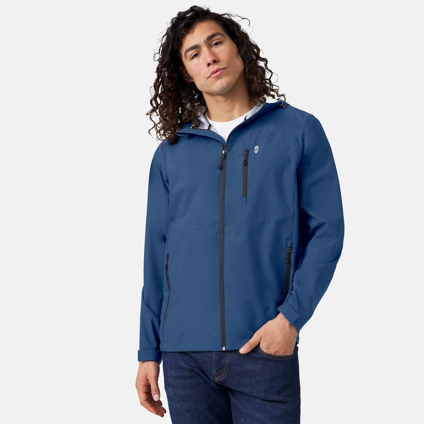 A person with long, curly hair wears the Clearance Men's Hydro Lite X2O Highline Waterproof Jacket in blue, featuring three zippered pockets, paired with dark jeans against a plain light background.