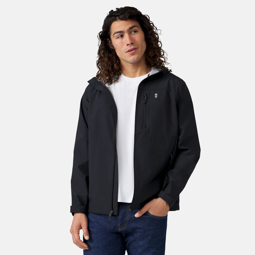 A person with long curly hair wears the Clearance Men's Hydro Lite X2O Highline Waterproof Jacket in black over a white t-shirt and blue jeans, standing with one hand in their pocket against a plain light background.