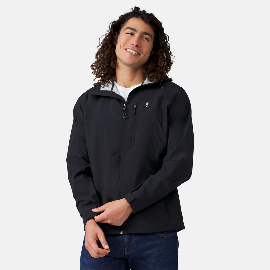 A man with long curly hair smiles while wearing the Clearance Men's Hydro Lite X2O Highline Waterproof Jacket over a white shirt and blue jeans, standing against a plain light gray background.