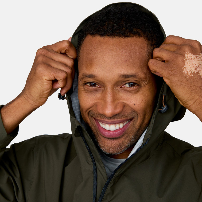 A smiling man with short hair and vitiligo on his right hand adjusts the hood of a Clearance Men's Hydro Lite X2O Highline Waterproof Jacket in olive-green, looking directly at the camera against a plain background.
