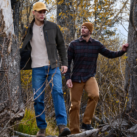 Two men walking outdoors among trees and brush in autumn foliage, wearing outdoor apparel. One wears a beige henley shirt, dark olive zip jacket, blue jeans, and a tan cap.