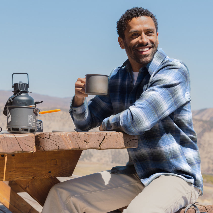 Man sitting outdoors holding a coffee mug with a clear blue sky and mountains in the background
