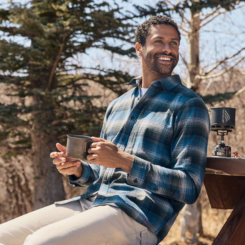 Man in a plaid shirt holding a mug outdoors with trees in the background