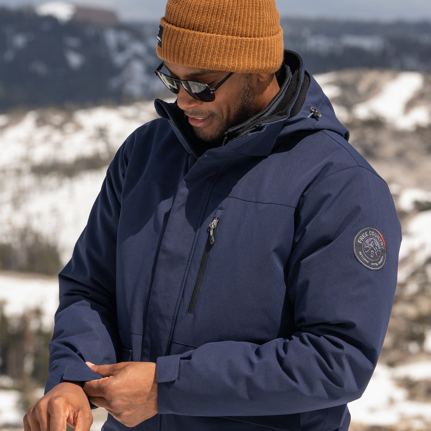 Man wearing Free Country Men's Ski Patrol 3-in-1 Systems Jacket in dark navy with a full front zipper opening, an outer jacket for wind and cold weather, an inner puffer jacket, and wind resistant and water resistant stretch performance fabric, light grey background. Perfect menās jacket for winter weather.