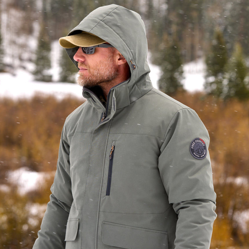 Man wearing Free Country Men's Ski Patrol 3-in-1 Systems Jacket in grey with a full front zipper opening, an outer jacket for wind and cold weather, an inner puffer jacket, and wind resistant and water resistant stretch performance fabric, light grey background. Perfect menās jacket for winter weather.