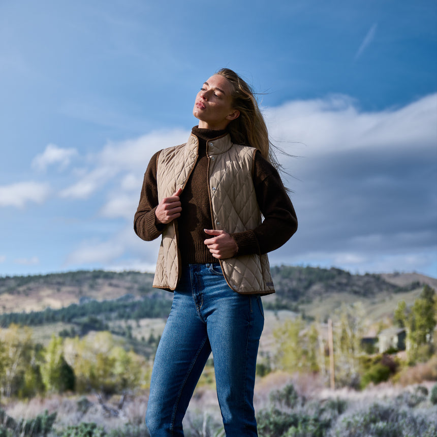 A woman stands outdoors in a field, wearing a brown sweater, blue jeans, and the Women’s Cirrus Diamond Quilted Button Down Vest by Free Country. With her eyes closed and facing the sun, she is surrounded by hills and a blue sky in the background.